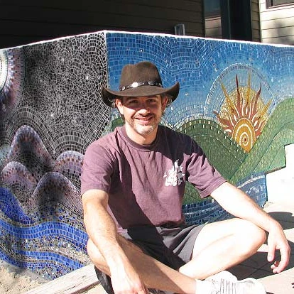 Shak, smiling in a brown hat, sitting in front of a colourful mosaic wall